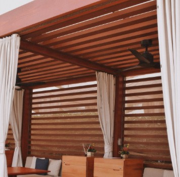 Pérgola de lamas con cortinas para terraza Pérgola de lamas de madera con cortinas laterales y ventilador en un porche
